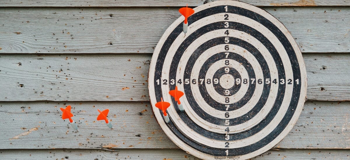 An image of a target with a bullseye target against a plain wooden wall. The dartboard has darts all aound it but none hit the target, implying failure