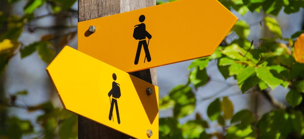 An orange arrow sign and a yellow arrow sign, both with black images of backpackers, pointing in two different directions, implying uncertainty, geopolitical or otherwise