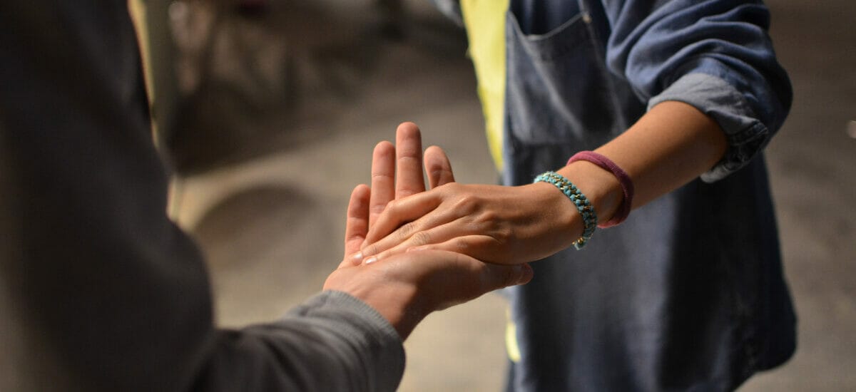 A man lending a woman a helping hand, leading her with their palms together. She is wearing a blue denim shirt and he has a dark gray suit jacket. You cannot see their faces - just their hands and midsections.