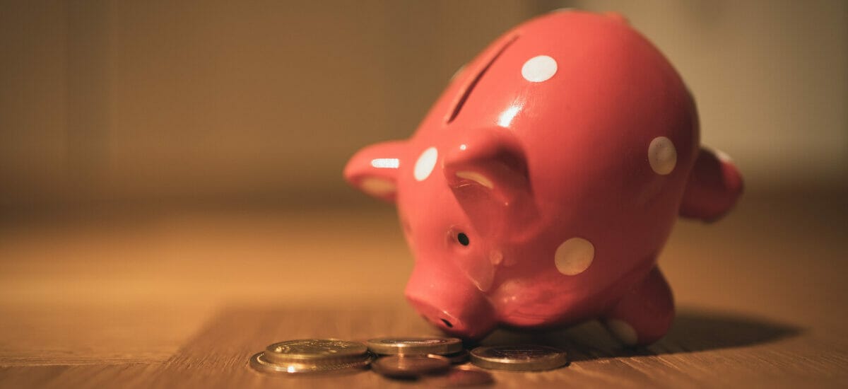 A pink ceramic piggy bank on a wooden table looking down nose-first at some scattered coins