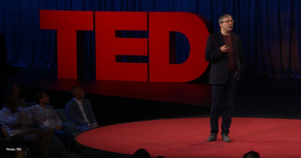 Gary Marcus presenting on stage at his TED Talk. He is wearing a dark jacket, dark jeans and a maroon shirt, standing on a red stage in front of large red letters that read "TED"