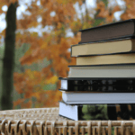 A photograph of a pile of fall books sitting on a wicker table with autumn trees in the background